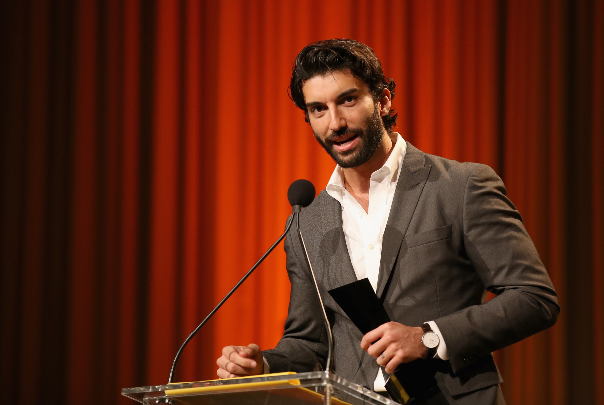 Actor Justin Baldoni speaks into a microphone, wearing a gray suit and white shirt, in front of a red-curtain backdrop.