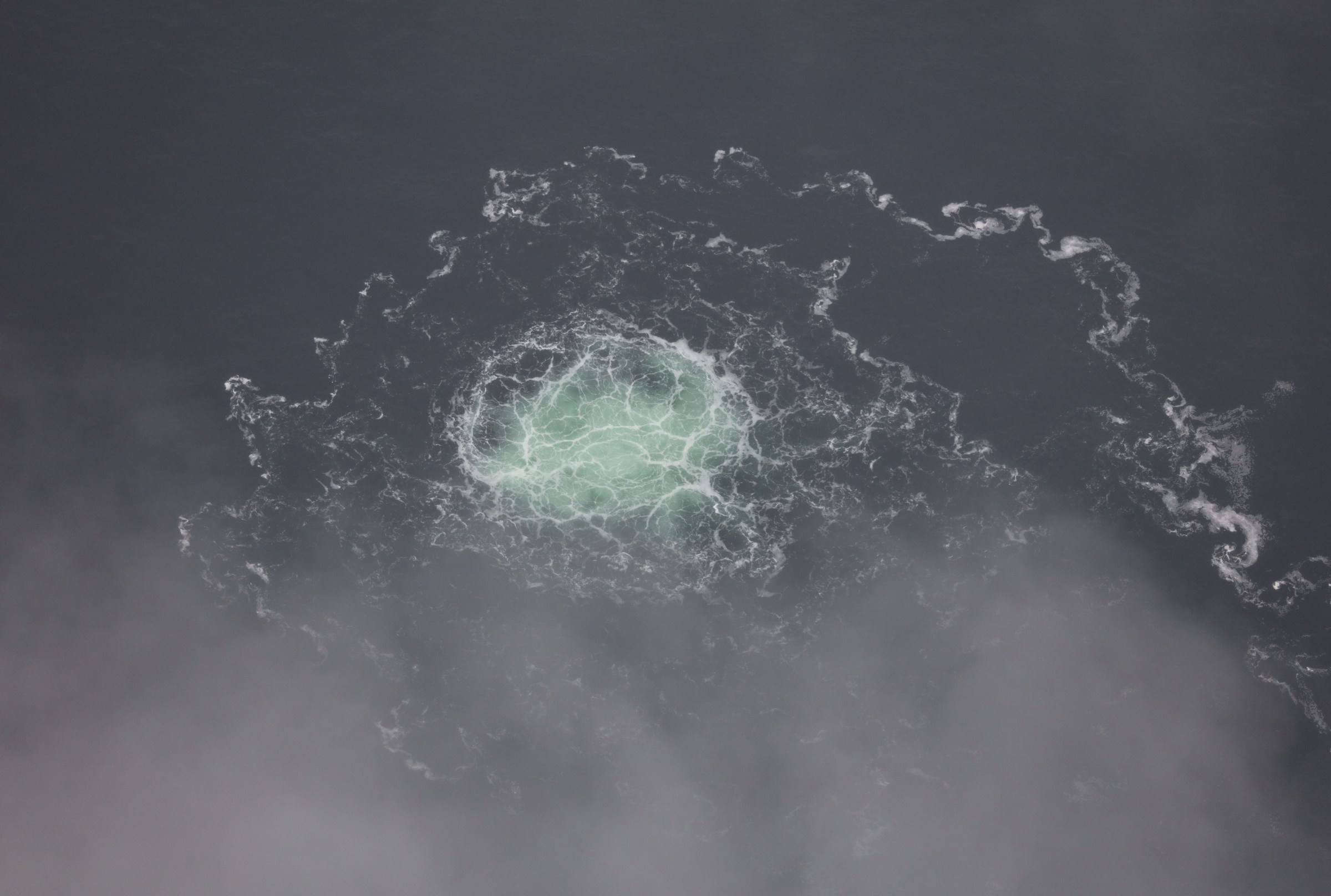 Greenish gas bubbles rise to the surface, with clouds of bubbles underneath, in a gray expanse of water.