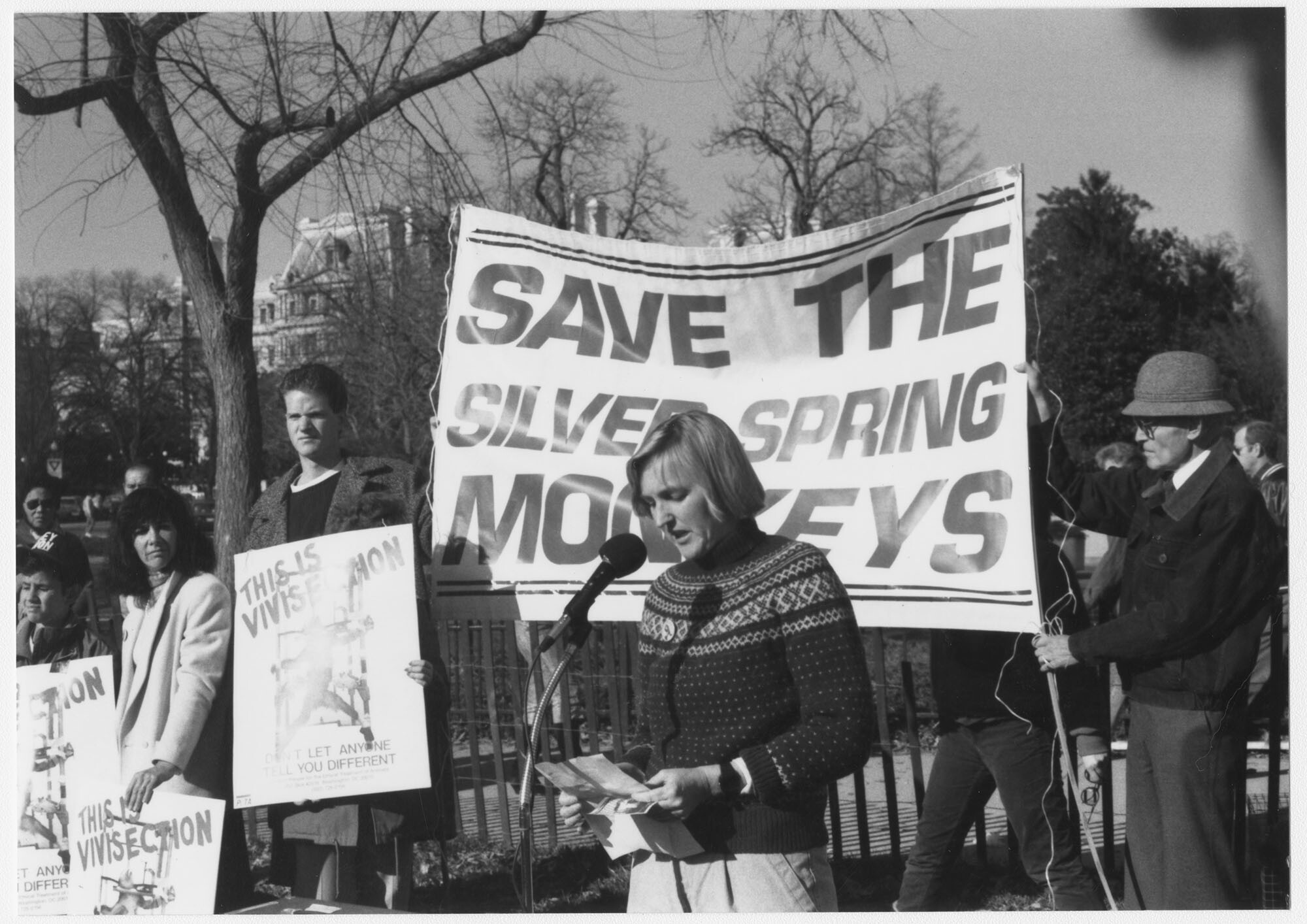 black-and-white photo of a crowd holding animal testing protest signs, a large banner reads “SAVE THE SILVER SPRING MONKEYS.” A blond woman stands in front of a mic speaking