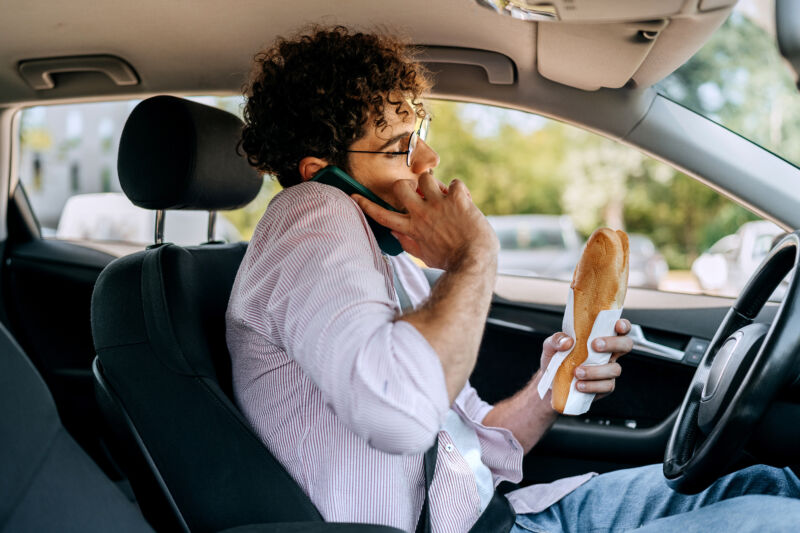 Young casually clothed man talking on a phone and eating sandwich while driving car