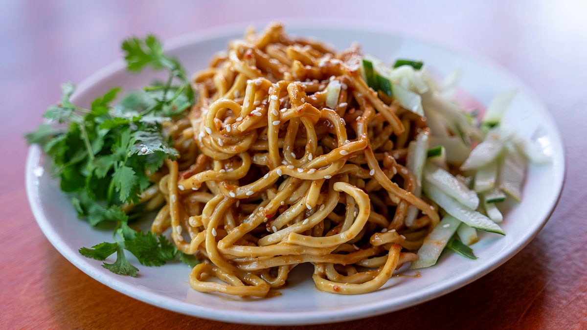 A plate of chilled egg noodles with cilantro and cucumber on the side. The noodles are tossed in a brown sauce and topped with sesame seeds.