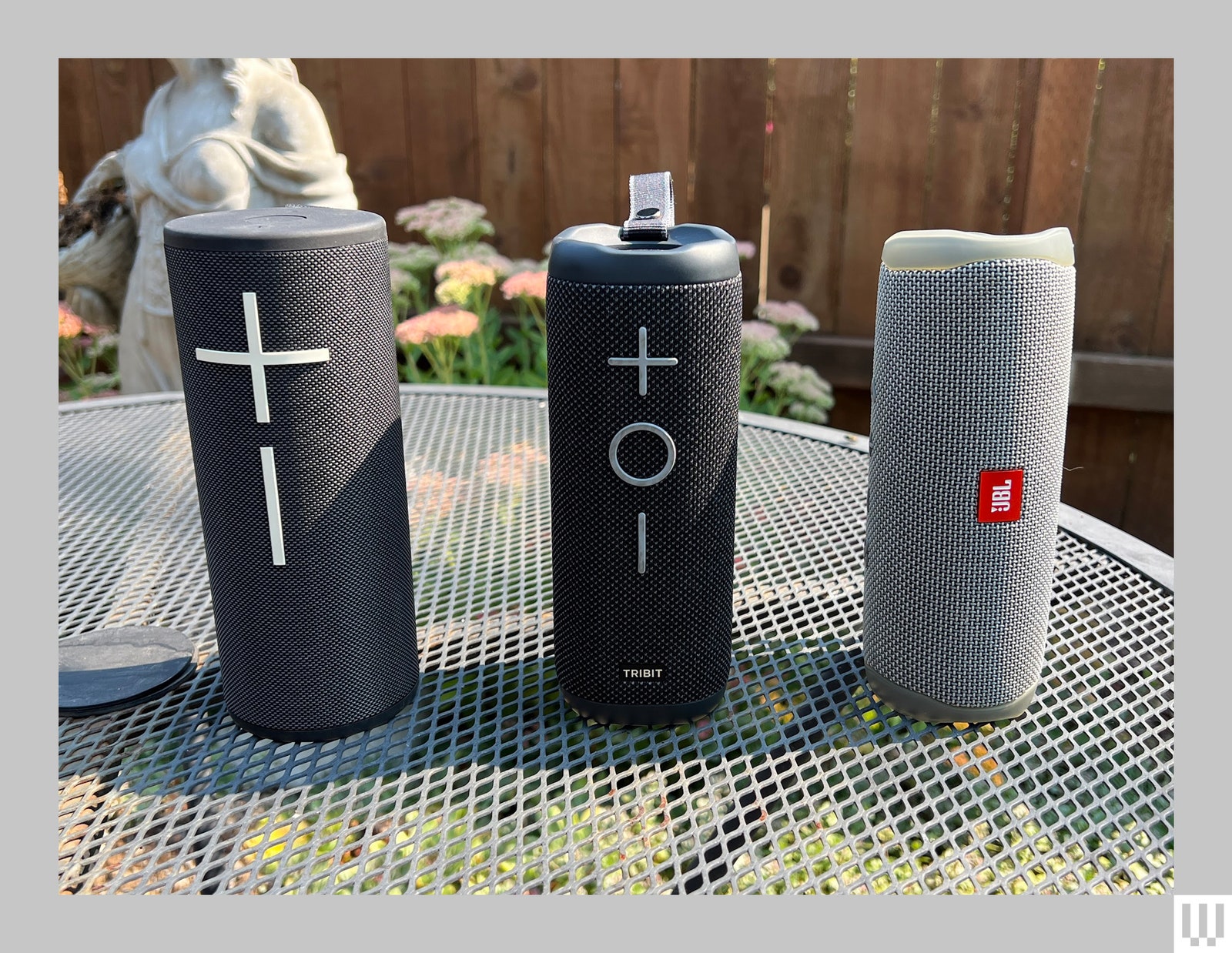 2 black and 1 grey cylindrical portable speakers sitting on a mesh metal garden table with a small sculpture and wooden...