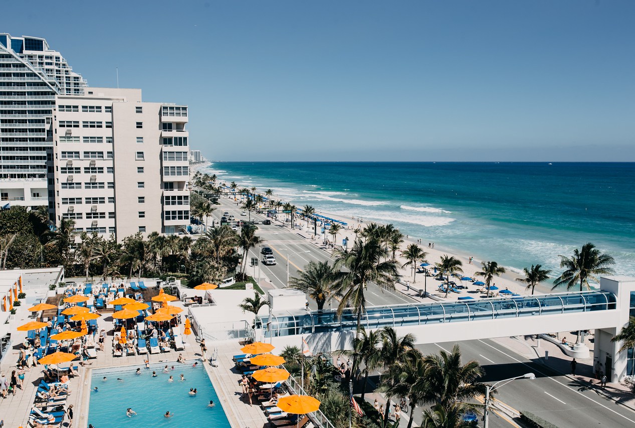 An aerial photo of the seashore in Miami, featuring a four-lane road parallel to the beach and a high-rise hotel and pool opposite.