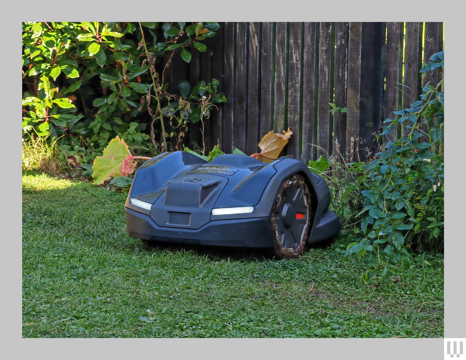 Side view of the Husqvarna robot mower showing it's black angular case and large wheels with a wooden fence and...