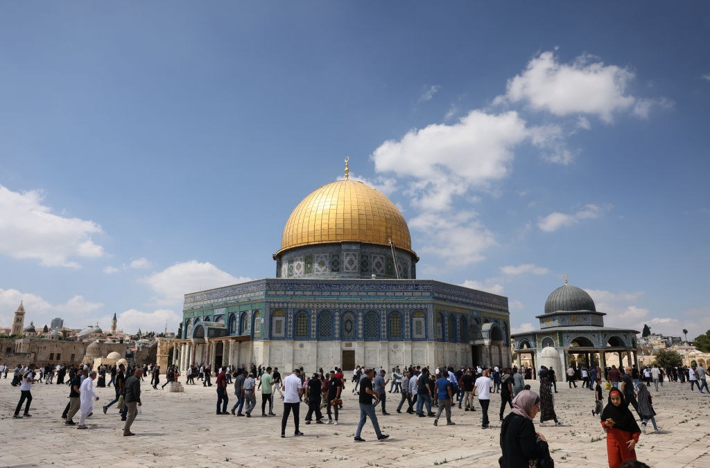 An ornate octagonal structure with blue Islamic mosaic walls and a golden dome. Crowds mill around on the stone square in front of the building.