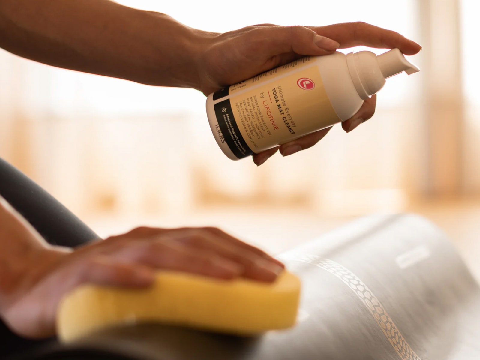 Hand holding a spray bottle aimed toward a rolled up yoga mat while the other hand wipes the mat with a sponge