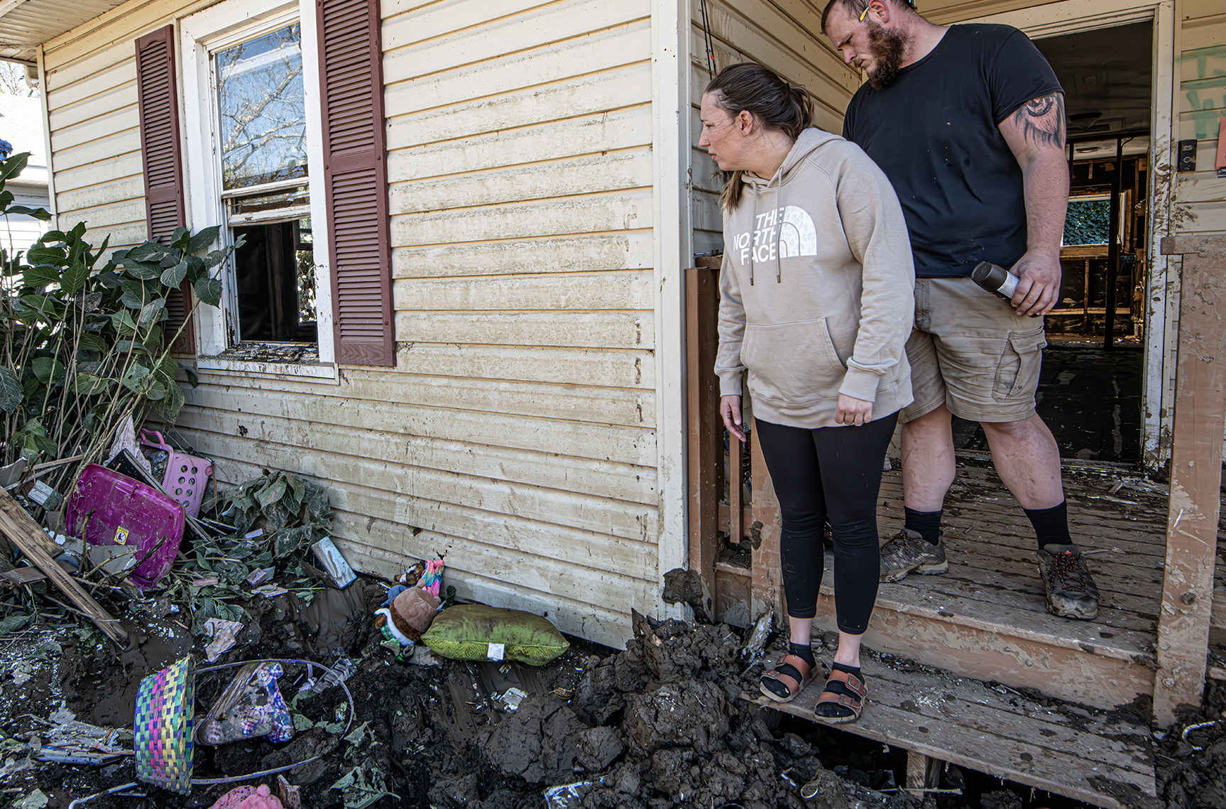 A man and a woman stand on their front porch looking out on their personal belongings scattered in their front yard.