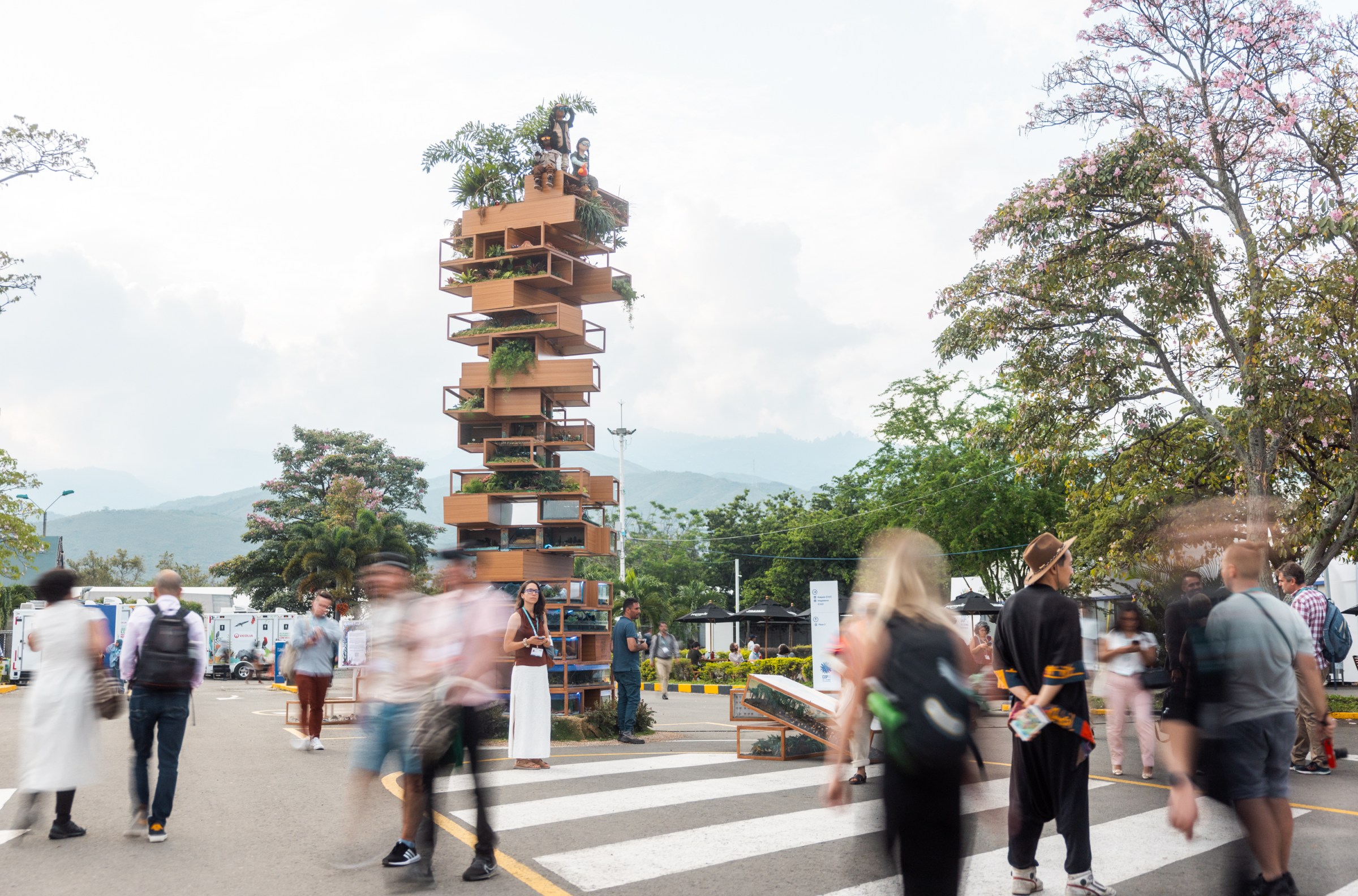 People walk across a paved outdoor space surrounded by trees, with hazy hills in the distance. At the center of the space is a sculpture that looks like a Jenga tower with pieces missing, with plants cascading out of some blocks.