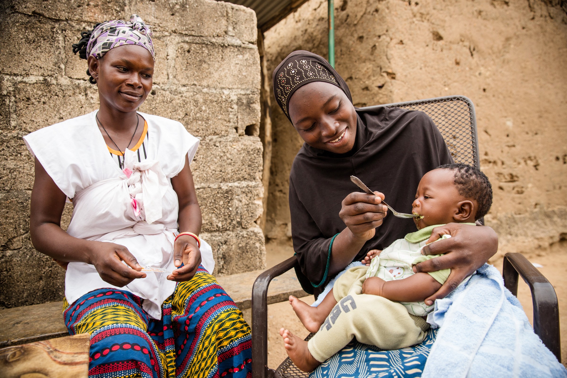 A health worker looks on as a mother spoon-feeds the baby on her lap.