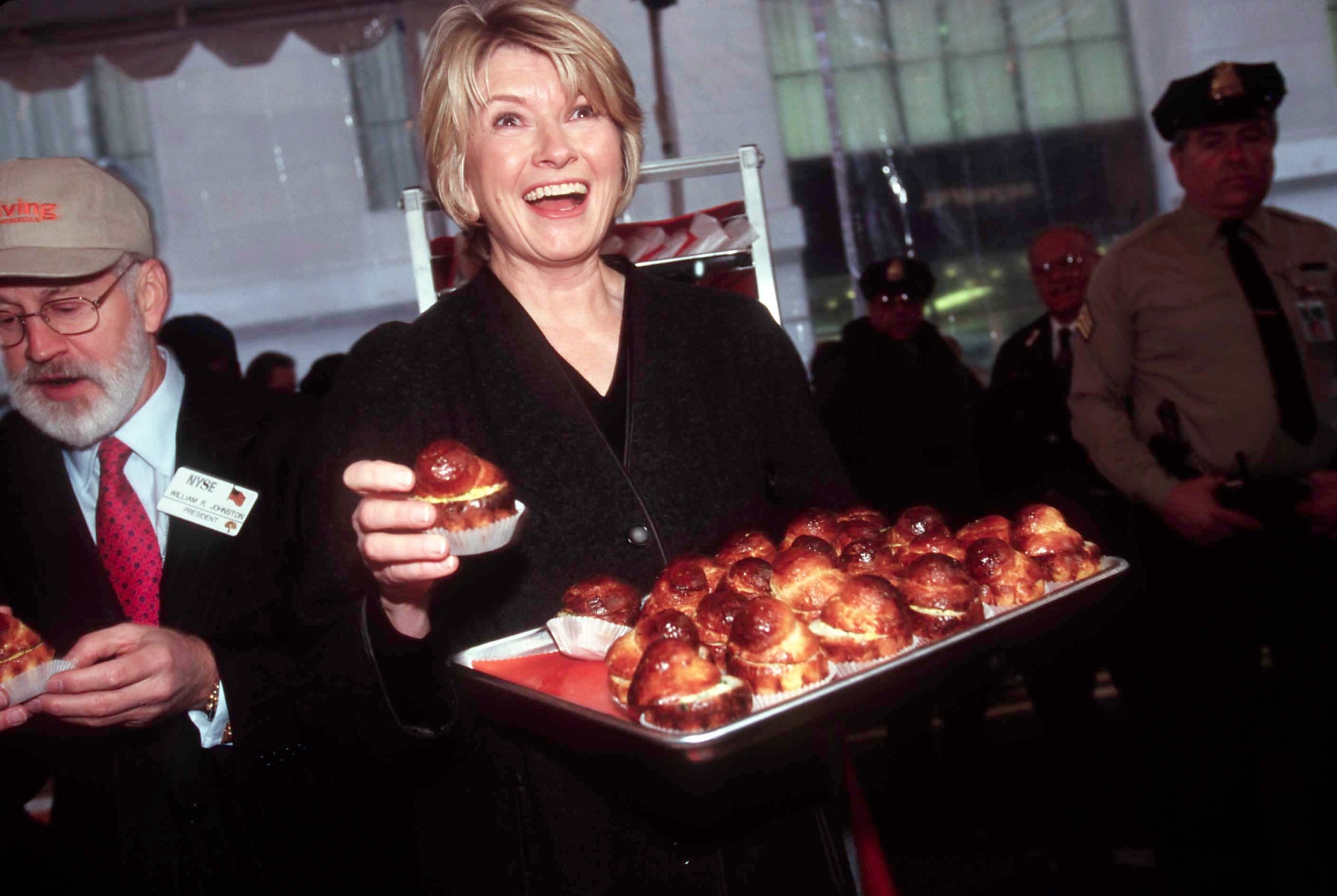 Martha Stewart, beaming, carries a tray of brioche sandwiches through the glass doors of the Stock Exchange. To her right an older man with a white beard has started to eat one of the sandwiches.