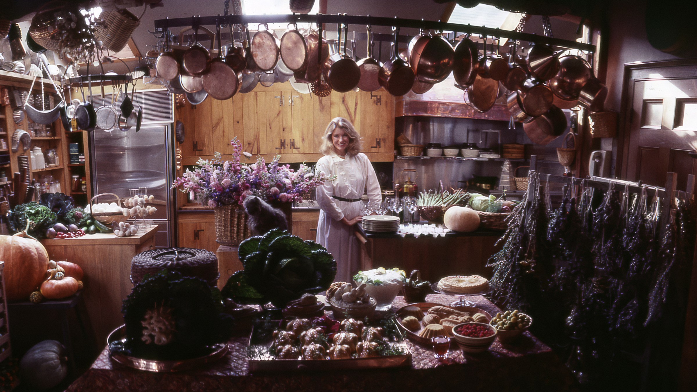 A blonde woman in her 30s stands in the center of a cluttered wood-paneled kitchen. Over her head hang dozens of copper pots and several stainless steel utensils. On the countertops around her are dried flowers, pies, cracker plates, pumpkins, asparagus, other vegetables, and a cat.