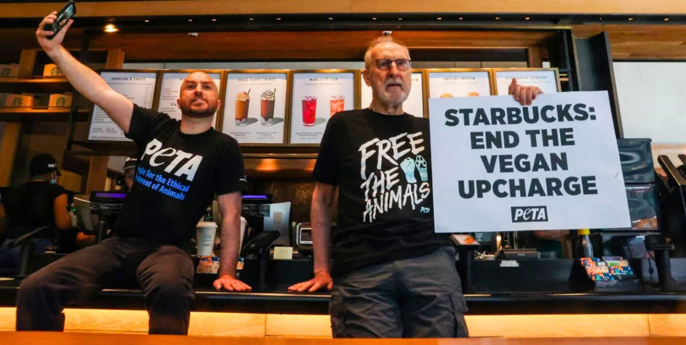 Two men wearing PETA shirts protest at a Starbucks counter, one holds a sign that reads “STARBUCKS: END THE VEGAN UPCHARGE”