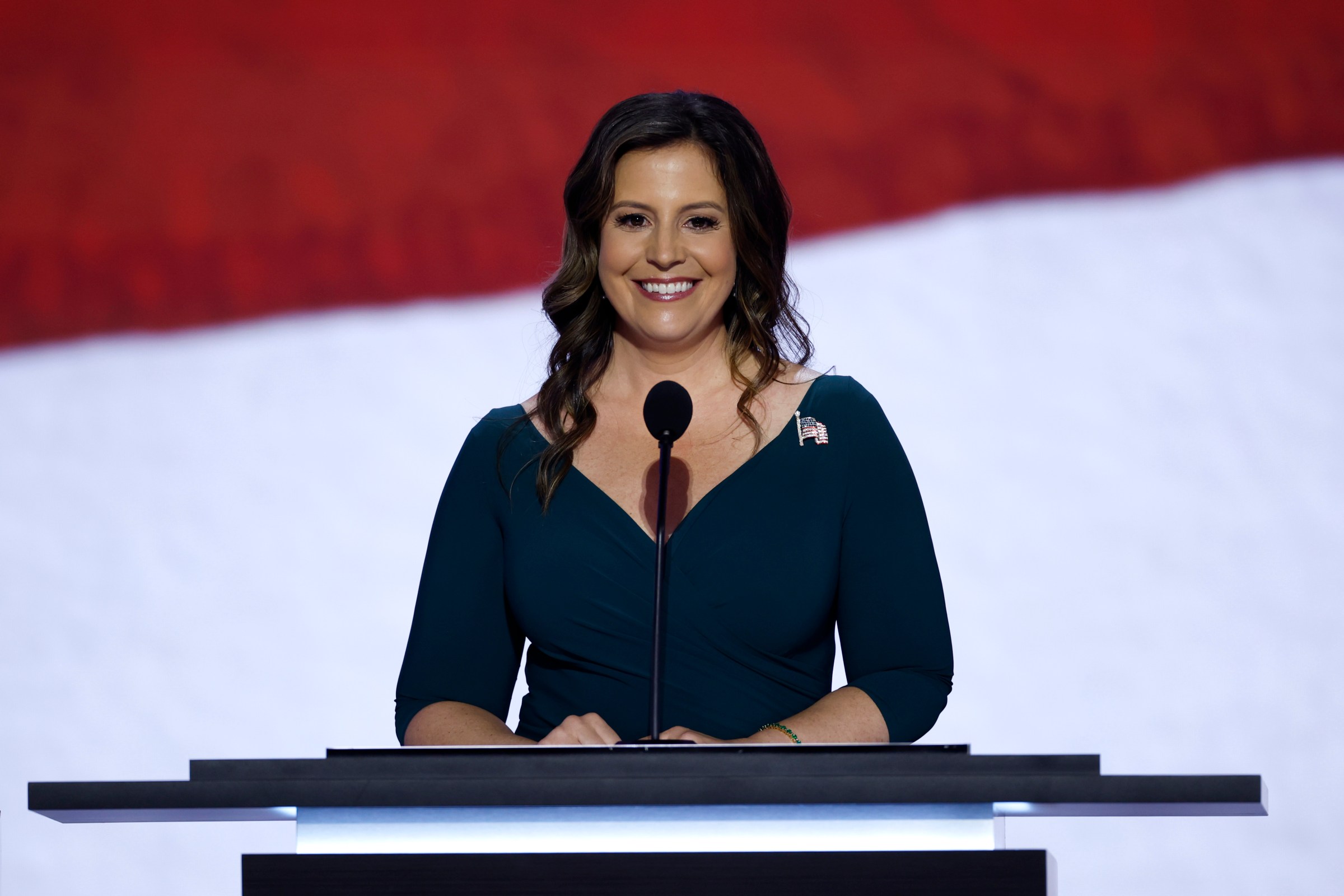 Representative Elise Stefanik in a dark blue V-neck dress, standing and smiling in front of a microphone.