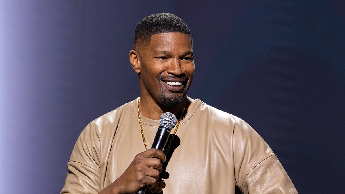 Comedian Jamie Foxx holds microphone while wearing brown leather shirt.