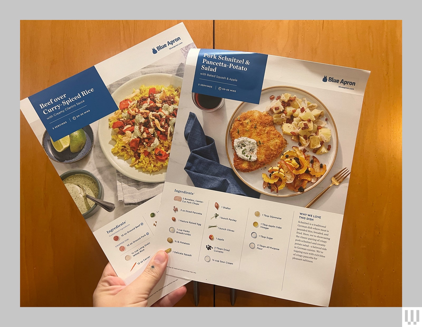 A hand holding two large recipe cards from Blue Apron a delivery meal kit