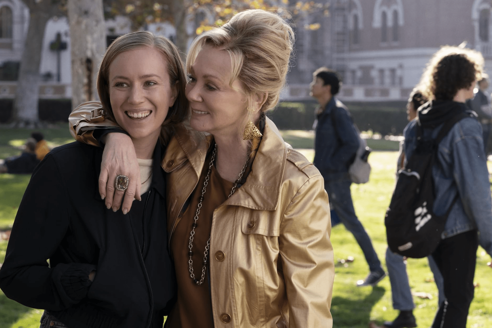 Two blond women in a park stand close and smile.