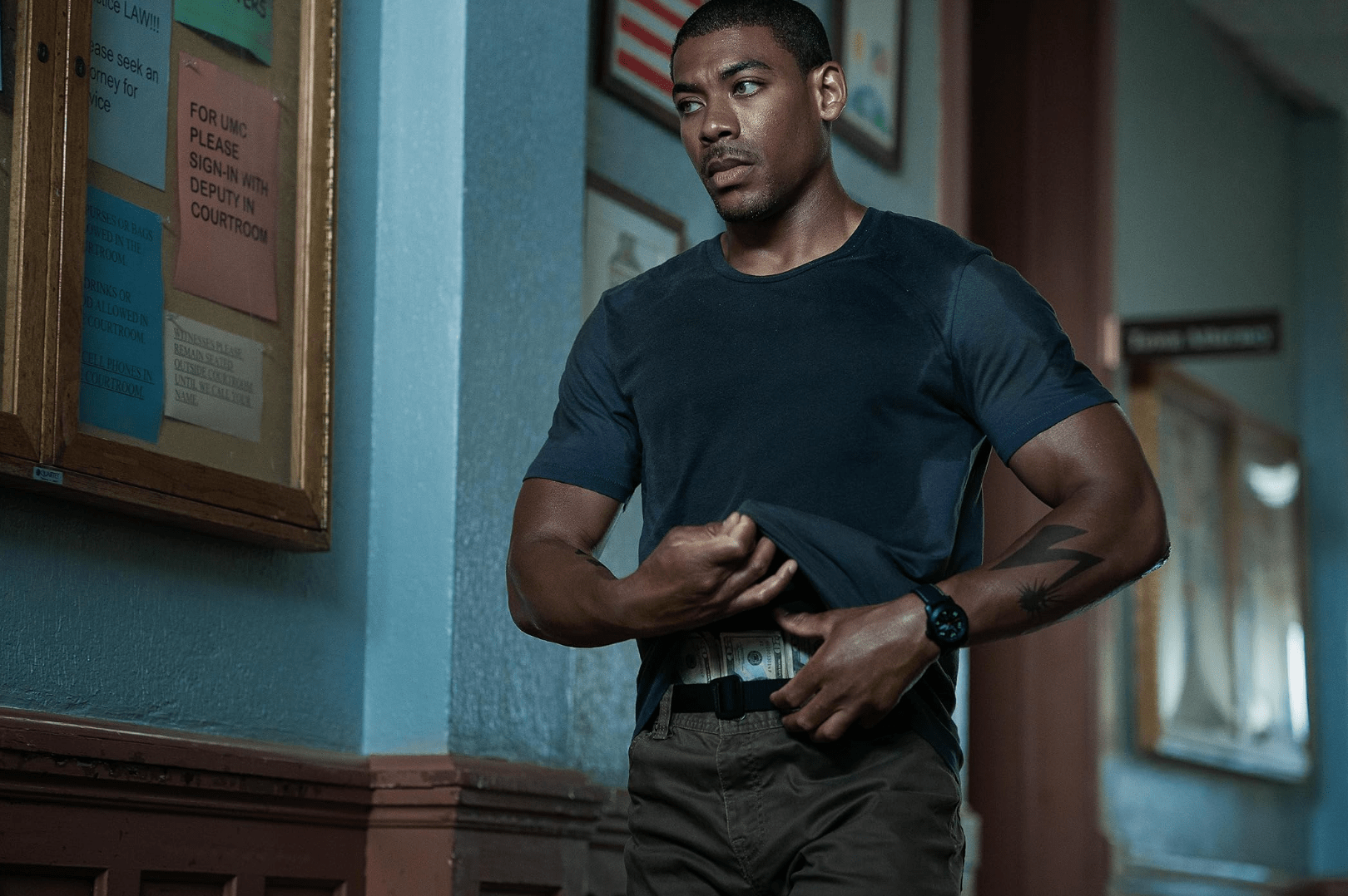 Aaron Pierre in a blue shirt with cash in his waistband.