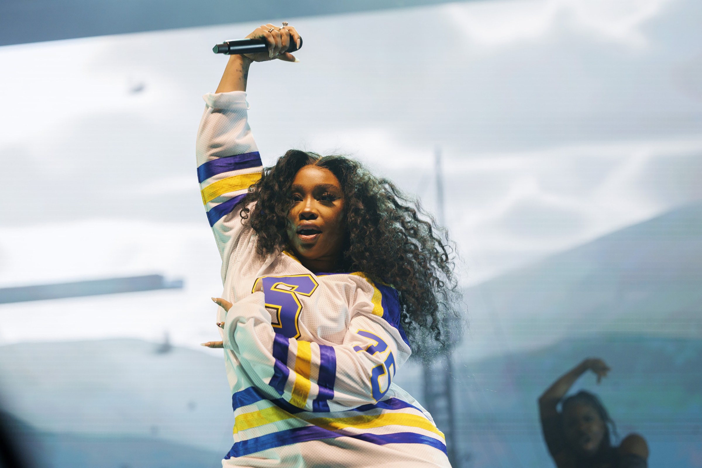A young Black woman with long hair and wearing a white, blue, and yellow football jersey dances onstage with a microphone held over her head.