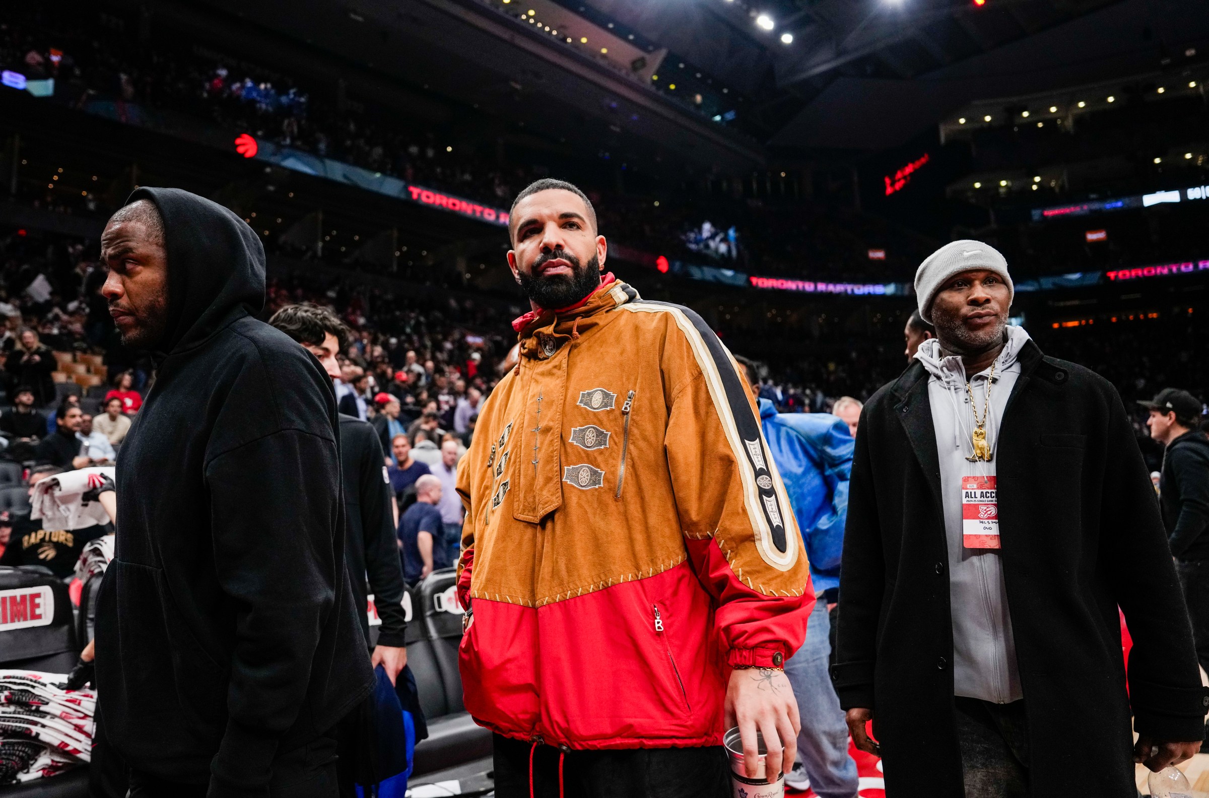 Drake wears an orange and red windbreaker with his bodyguards at a basketball game