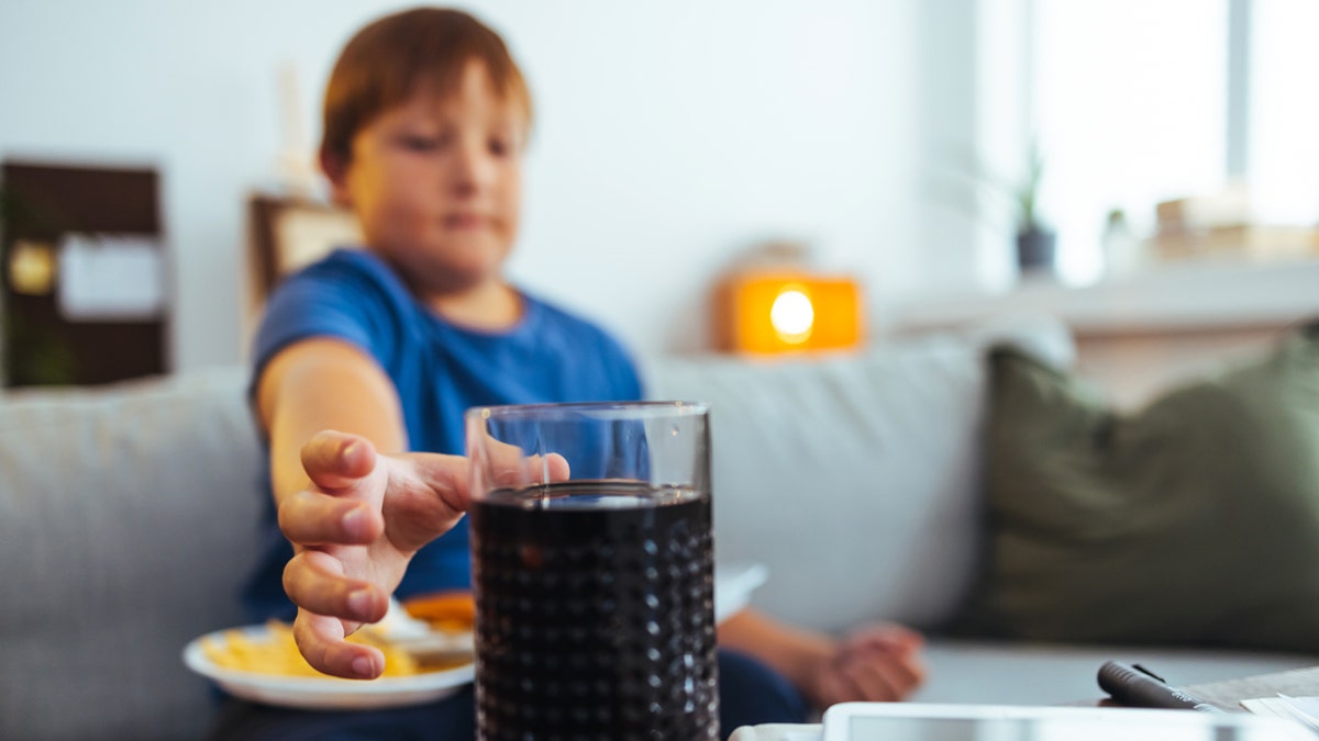 An obese boy reaches for a glass of soda.