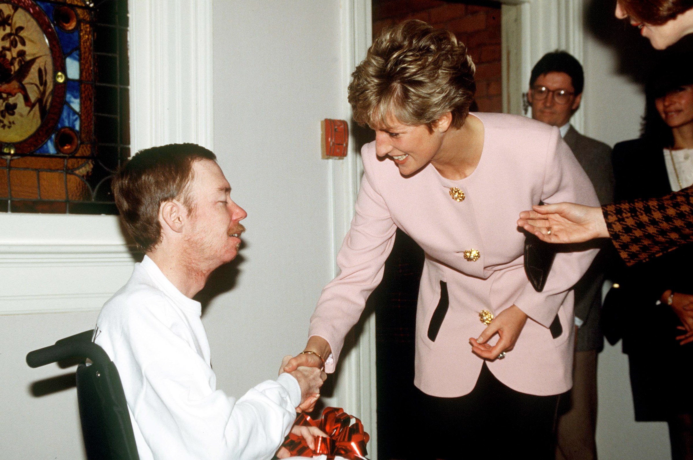 Diana, in a pink and black jacketed suit, shakes hands with a frail man in a wheelchair, while other people stand behind her.