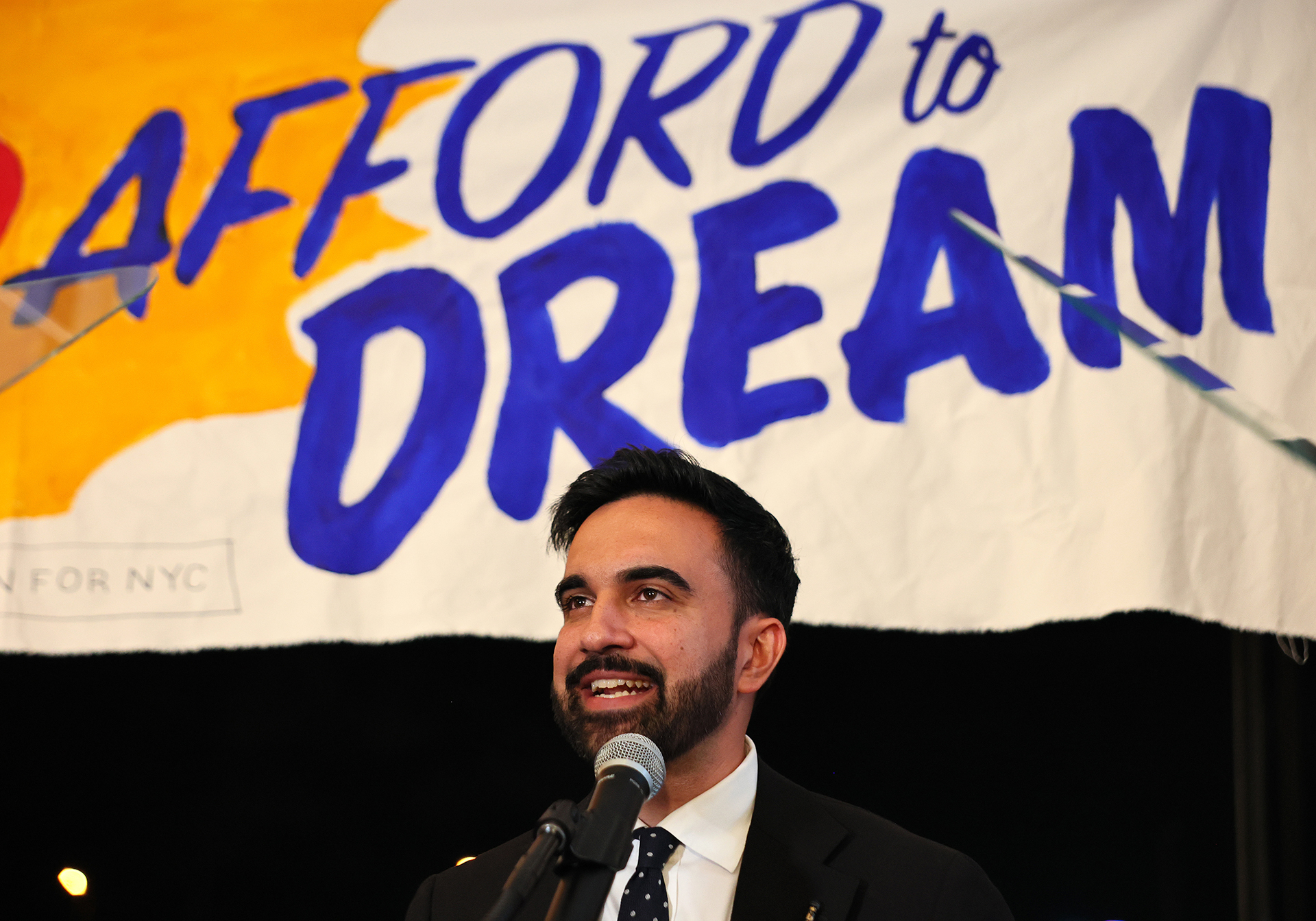 Zohran Mamdani speaks in front of a poster that reads “Afford to Dream”