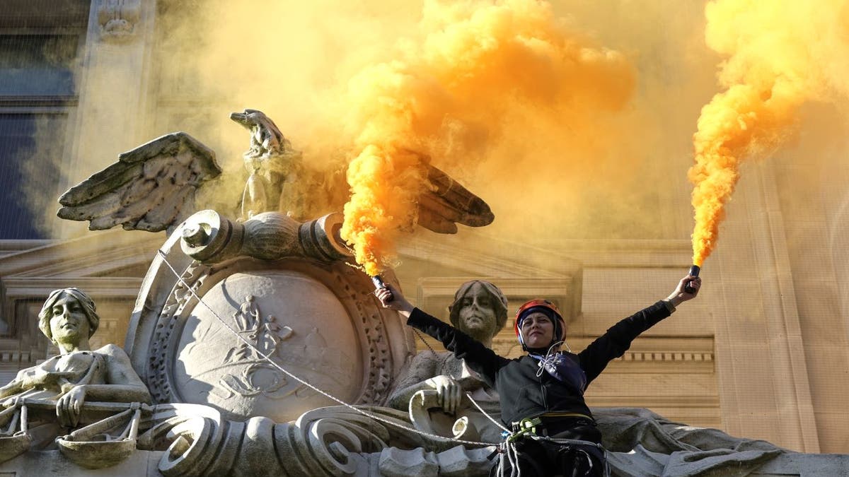 Extinction Rebellion activist climbs DC building with orange smoke during fossil fuel protest.