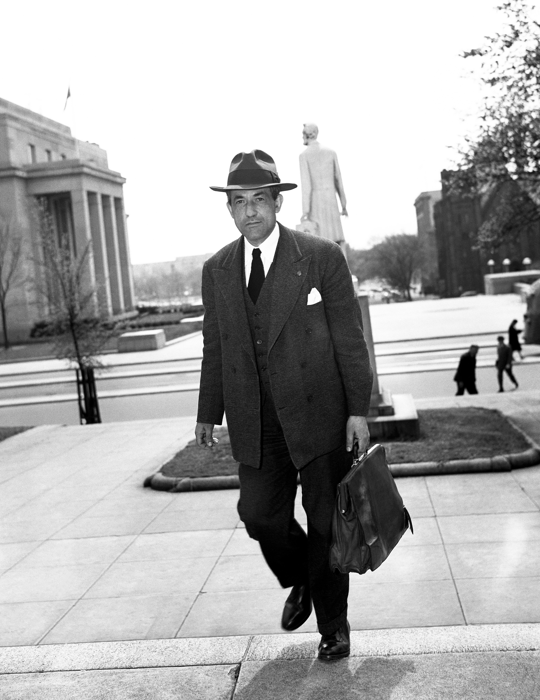 A black and white photo of Lawrence Dennis walking up steps in a suit with a briefcase.