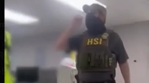 X A masked man wearing a khaki green police vest with HSI in yellow written on the front. He has a police badge pinned to the vest shoulder and is wearing a dark T-shirt. 