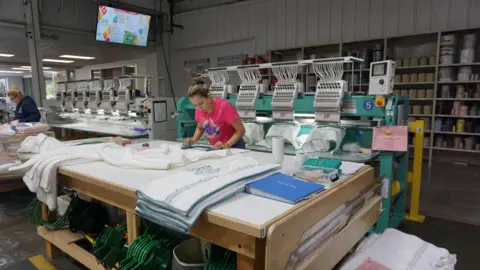 A worker in a pink shirt makes towels at the Matouk factory in Fall River, Massachusetts. She is examining a white towel on a large workbench, standing in front of a large teal green machine that appears to be embroidering patterns onto other towels.
