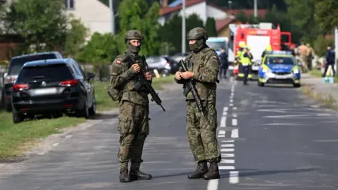 EPA Two members of the army stand on a street, in uniform holding guns, as emergency services gather in the background. 