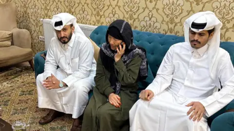 Reuters Faye Hall sits on a blue sofa in between two men wearing white in a plush room with patterned yellow wallpaper and carpeting. She wears a long dark green dress with long sleeves and a black and white head scarf and is speaking into a mobile phone.