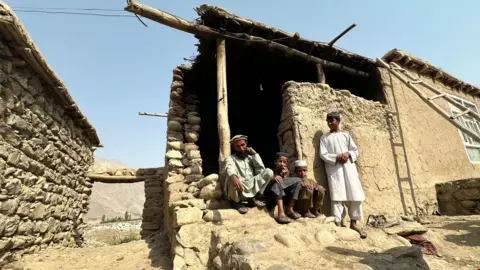 Aakriti Thapar / BBC Khan Mohammad is pictured sitting outside a building with his family against a blue sky in the village of Shesh Pol.