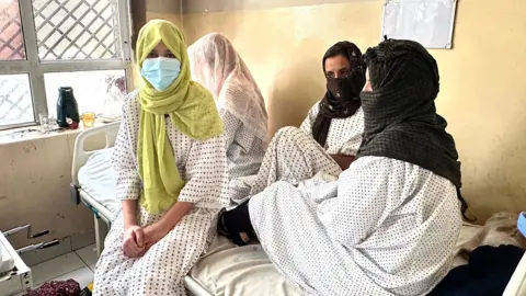 Aakriti Thapar / BBC Zuhra Shewan wears a green head scarf and medical mouth covering while sat on a hospital bed with three other patients.