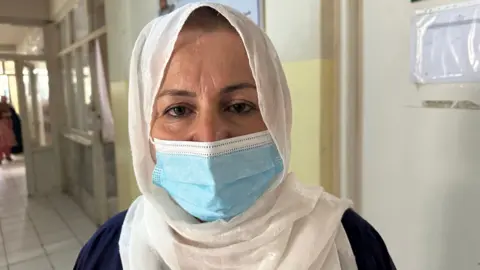 Aakriti Thapar / BBC A head-and-shoulders portrait of Razia Hanifi wearing a white head scarf and medical mouth covering in a corridor on the hospital's ward.