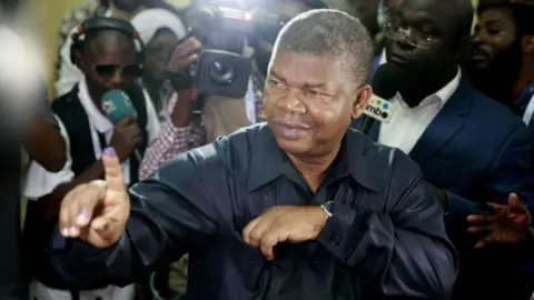 AFP via Getty Images Joao Lourenco holding an inked finger up to the cameras after he voted. He is surrounded by journalists and is wearing a black open-necked shirt.