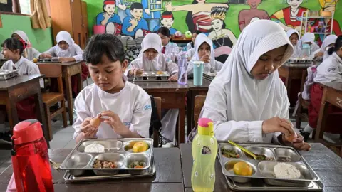 Getty Images Two young girls eat from metal trays at a classroom in Indonesia, with their classmates doing the same in the background.