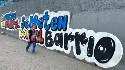 Nicole Kolster/BBC A woman wearing blue trousers and a black T-shirt walks past a grey wall with colourful graffiti reading reads: 'If you mess with Maduro, you mess with the neighbourhood"