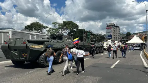 Nicole Kolster/BBC People walk past armoured vehicles in a street in the capital, Caracas. In the background, tents can be seen. 