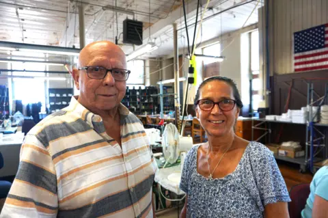 BBC Frank and Sue stand next to each other, smiling, in front of their factory floor. Behind them is a table with a fan, and an American flag hangs from the wall.