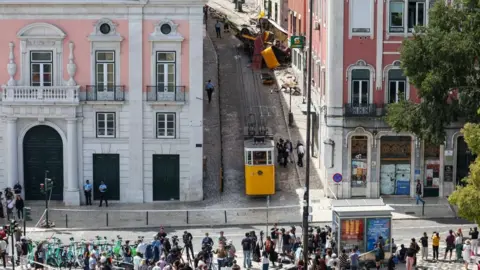 Reuters Several people, stood behind a group of television cameras look up a hill where the funicular crashed. 

The funicular in the accident is higher up the hill, damaged after the crash, and an intact once is at the bottom of the hill. A group of people stand next to the intact one, while two others are next to the damaged one.