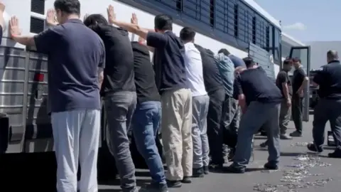 EPA/US Immigration and Customs Enforcement handout A still frame from a video shows a group of men, with their backs to the camera and hands on the side of a white coach with black bars on its windows during an immigration raid at the Hyundai-LG vehicle assembly plant in Ellabell, Georgia. 
The men are in casual clothing, mostly jeans or slacks and T-shirts. 