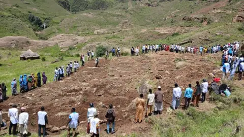Sudan Liberation Movement/Army People standing outside in a circle around a large muddy area which looks like a burial site 