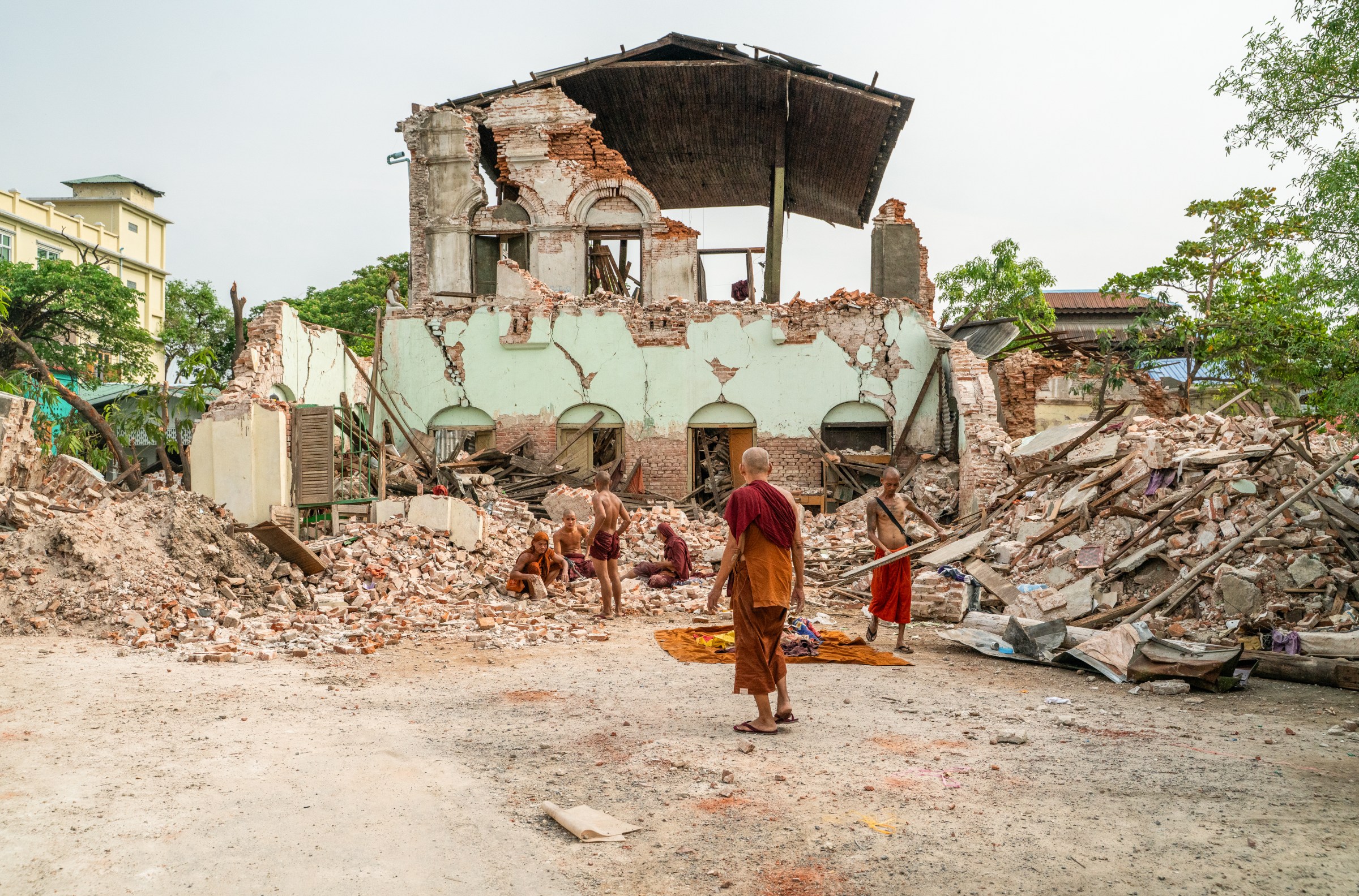 A number of monks stand in front of a collapsed building surrounded by rubble from other collapsed buildings around them.