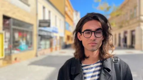 Ondrej Kapralek is a young man with shoulder length dark hair. He is wearing glasses with a black frame and a stripy t-shirt and blazer. He is standing in a pedestrian area with shops on either side. 