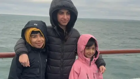 Three siblings stand together posing for a photo - Hussam on the left, Rahaf in the middle with her arms around her brother and Sara, on the right.  They are smiling and all have the hoods of their coats up. They are standing against the backdrop of the sea.