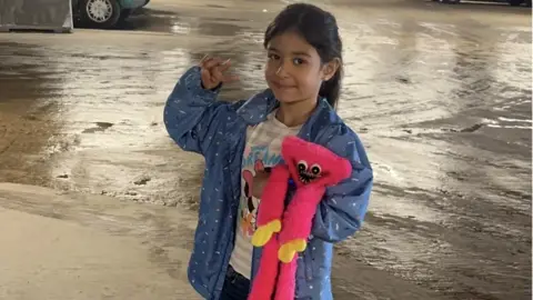 Sara stands smiling and doing a 'peace' sign with one hand, and holds a pink stuffed toy with the other. She wears a blue waterproof jacket and a top with Minnie Mouse on it underneath.  