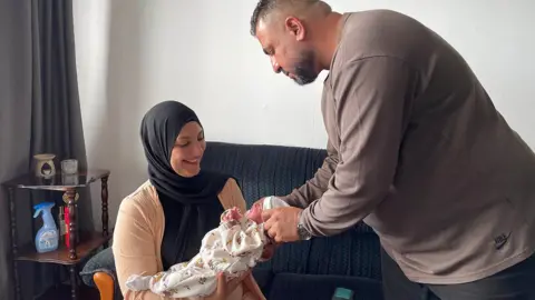 Nour sits on a dark coloured sofa, smiling as she cradles her newborn baby.  Her husband, Ahmed stands over them both and carefully adjusts the baby's white hat. 