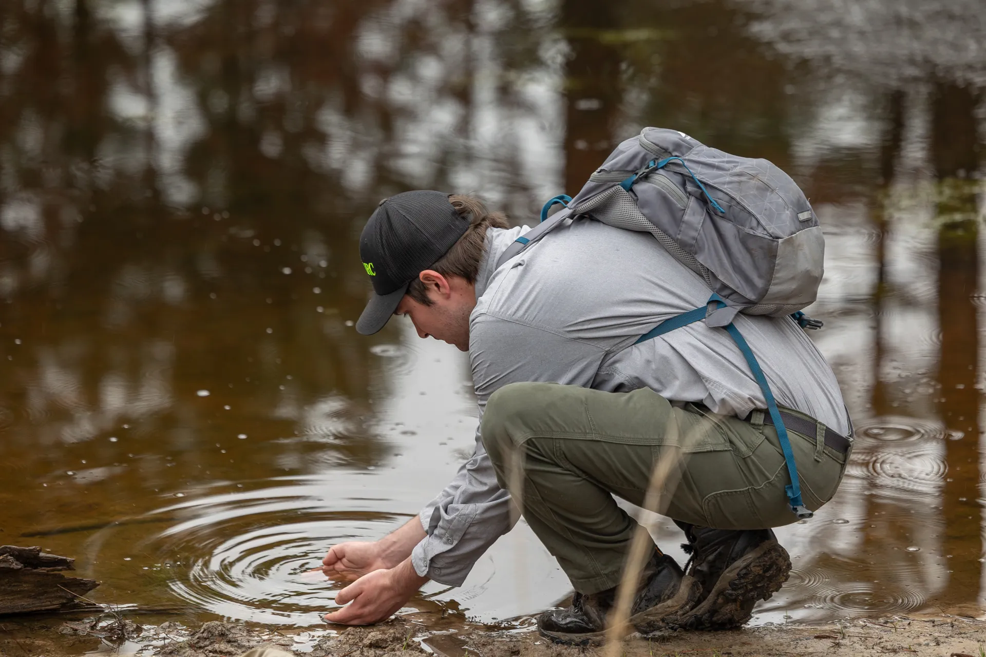 A man crouches down at the end of a pond, identifying tadpoles with his hands in the water