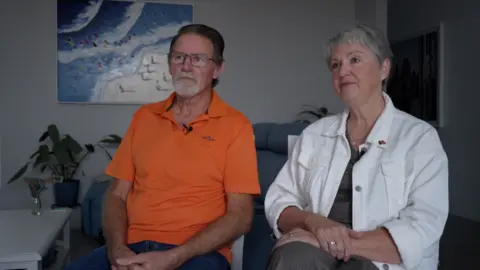 Dennis and Taryna Wawn who signed up for the VCL retirement cruise. They are sitting in their living room, he is wearing an orange polo shirt, and she is wearing a white shirt. A painting of a beach scene hangs on the wall behind them.